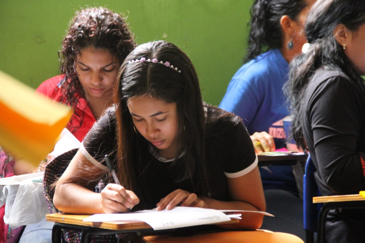 Joven sentada en un pupitre escribiendo y detrás de ella otros jóvenes.