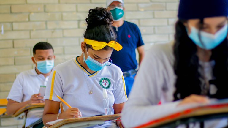 Jóvenes en sus pupitres escribiendo en salón de clases.