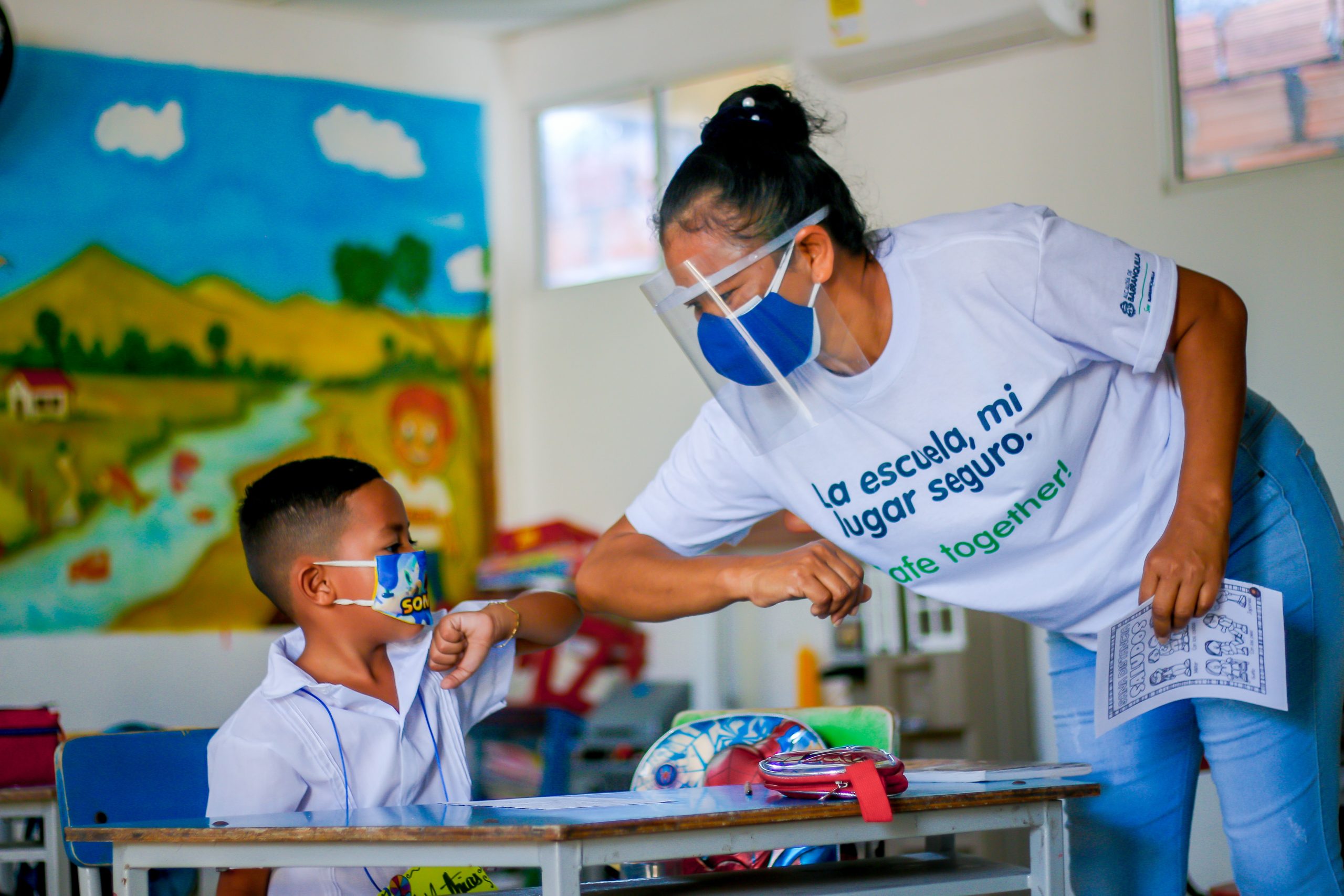 Profesora saludando con el codo a niño e el salón.