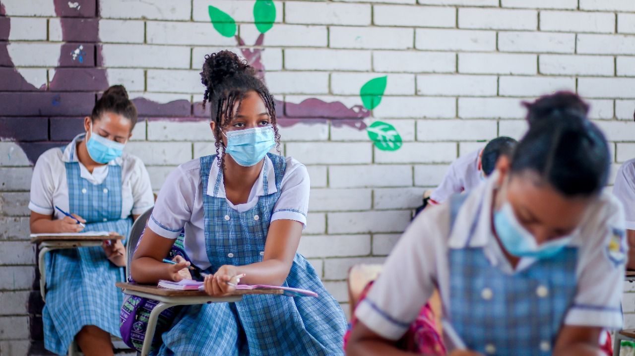 Adolescentes uniformadas escribiendo en sus pupitres en salón de clases.