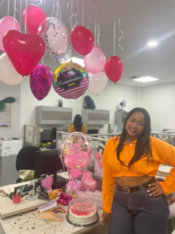 mujer recostada en su escritorio decorado con una torta, centro de mesa y globos colgantes en forma de corazón rojos y rosados