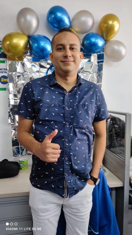 hombre con camisa azul recostado en su escritorio decorado con globos azules, dorados y plateados