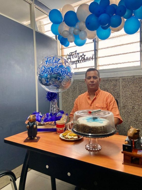 hombre sentado en escritorio con torta, centro de mesa de feliz cumpleaños y al fondo decoración de globos azules y blanco