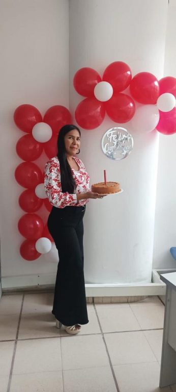 mujer de pie con torta de cumpleaños en las manos, al fondo decoración de globos rojos y blancos en forma de flores