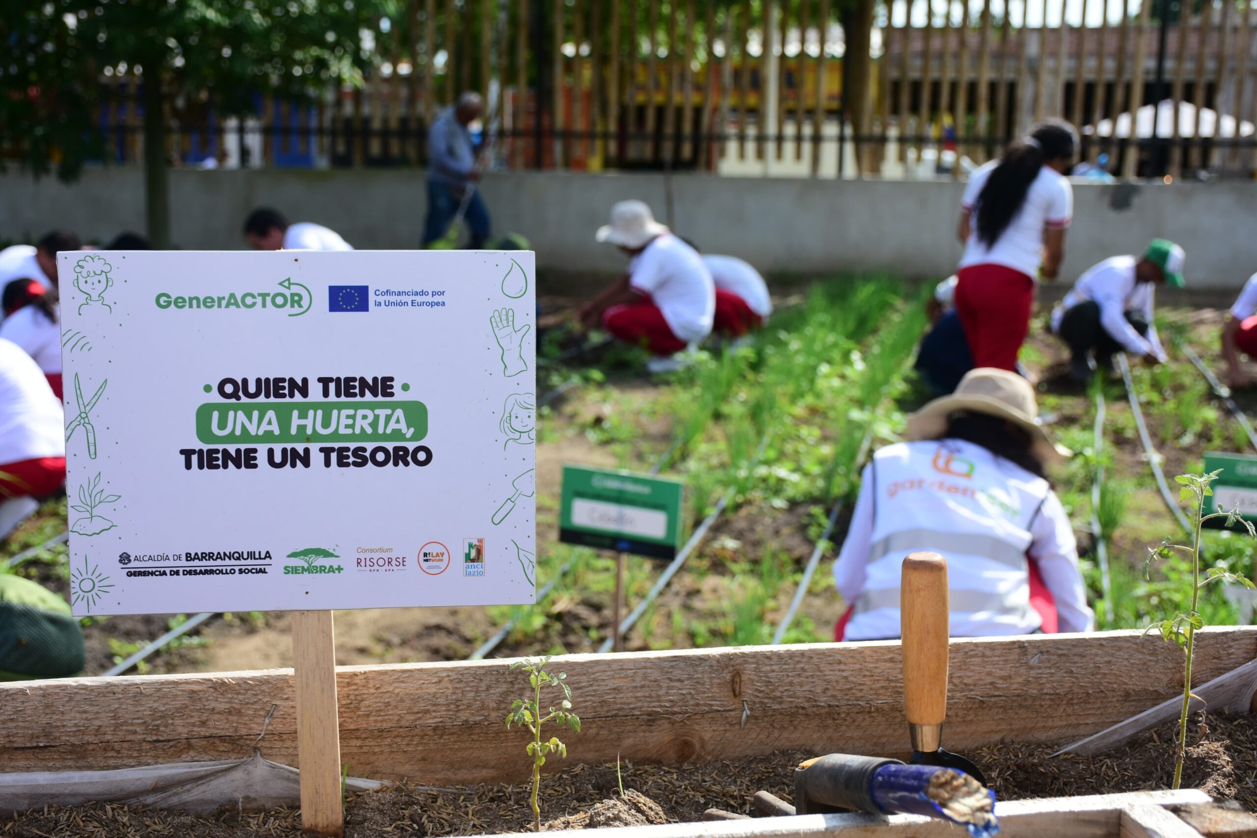 Personas cosechando en huerta Las Gardenias, junto cartel: "Quien tiene una huerta, tiene un tesoro".