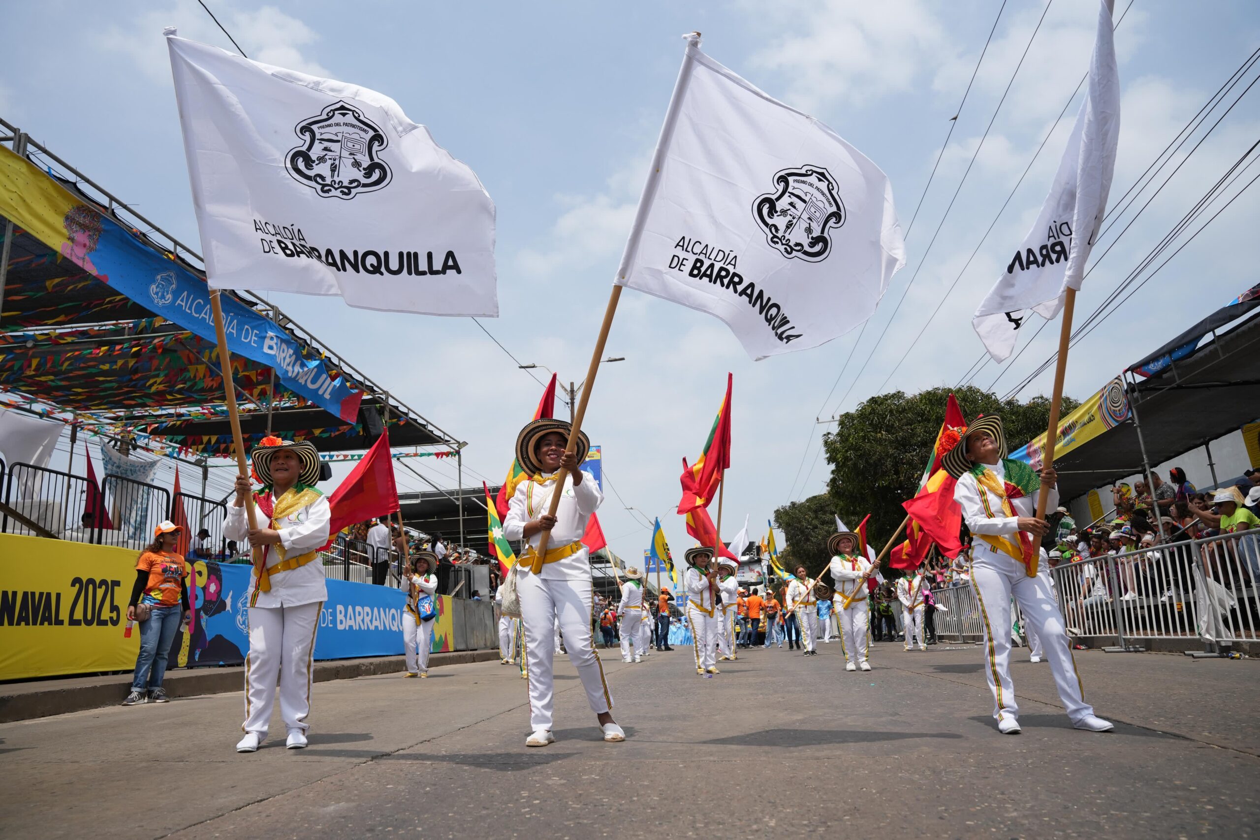 Grupo de personas vestidas de blanco sosteniendo banderas Alcaldía de Barranquilla en Carnaval 2025.