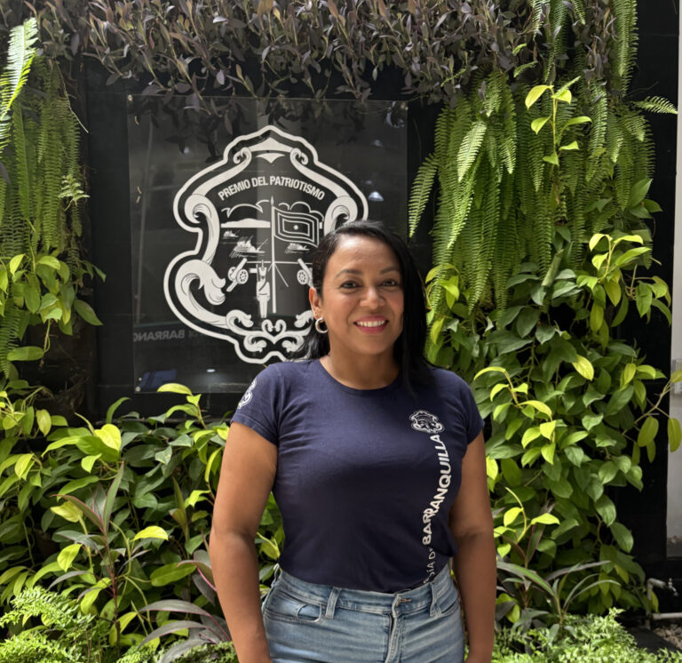Plano medio de funcionaria vestida de jean y camiseta institucional con un jardin vertical a sus espaldas.