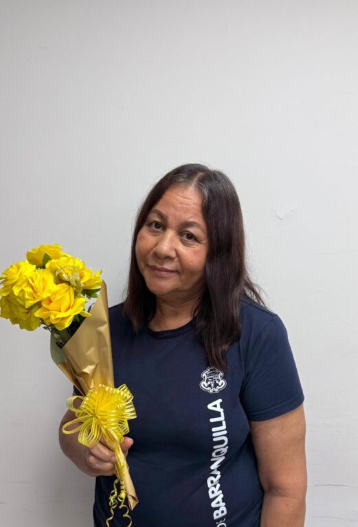 Funcionaria Leyla Cruz, en plano medio, con camiseta institucional color azul, sosteniendo un ramo de flores amarillas.