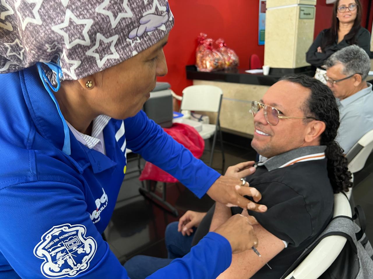 Ciudadano sonríe mientras recibe la vacuna contra la influenza por parte de una profesional de la salud.