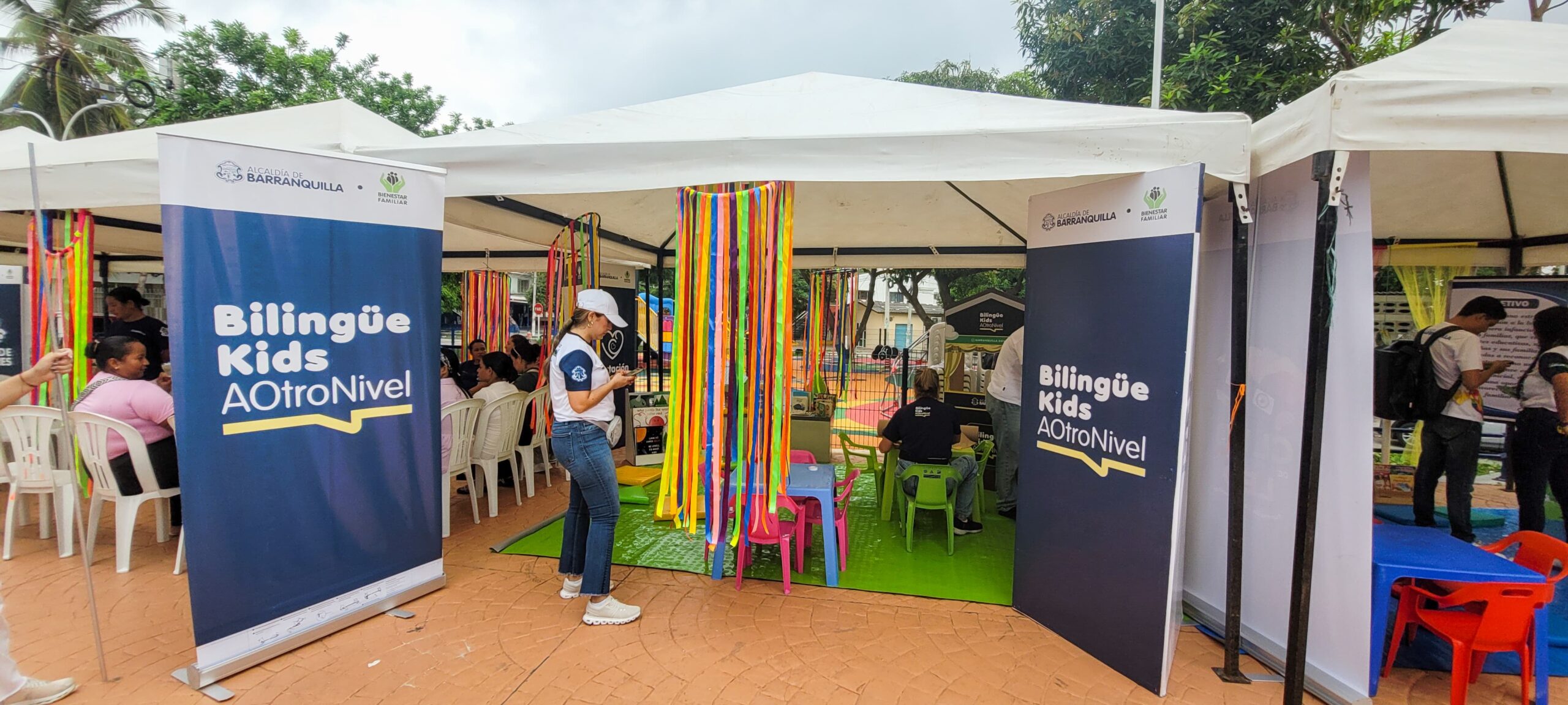 Carpa del programa “Bilingüe Kids A Otro Nivel” en un parque, con carteles, cintas de colores, mesas, sillas y material educativo infantil.