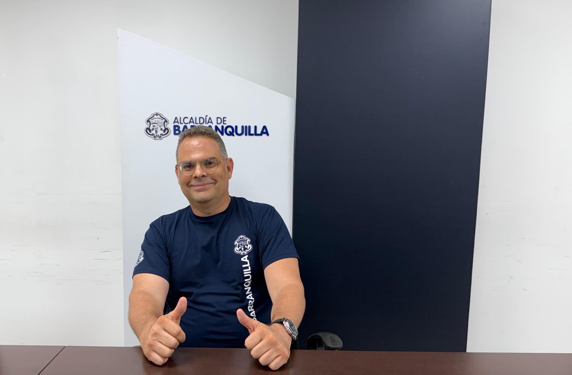 Funcionario Carlos Vergara, posa con uniforme institucional sentado frente a una mesa, con las manos con los dedos pulgares arriba, en señal de victoria.