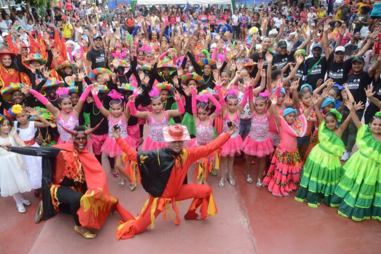 Grupo numeroso de niñas y niños en trajes típicos de carnaval, acompañados por adultos y personajes disfrazados.