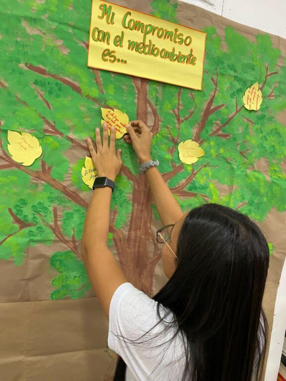 Una joven coloca una hoja con un mensaje en un árbol pintado en una cartelera para resaltar su compromiso con el medio ambiente.