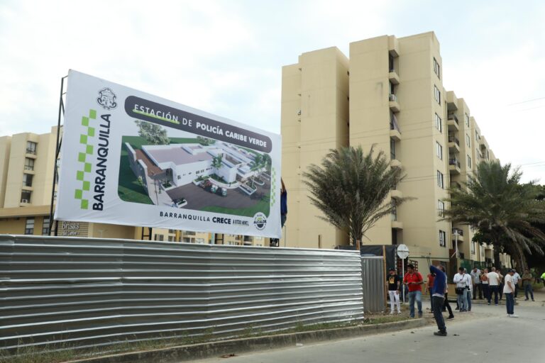 Aviso de obra anuncia futura Estación de Policía Caribe Verde en Barranquilla, con edificio residencial y personas alrededor.