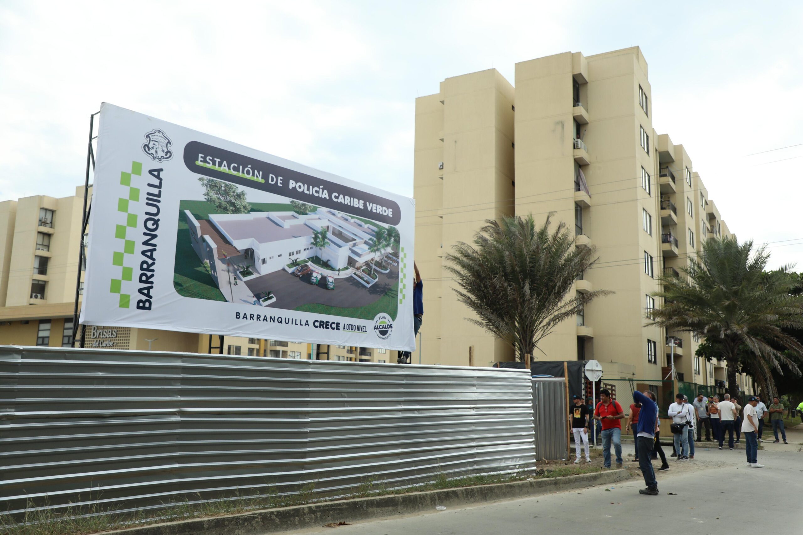 Aviso de obra anuncia futura Estación de Policía Caribe Verde en Barranquilla, con edificio residencial y personas alrededor.