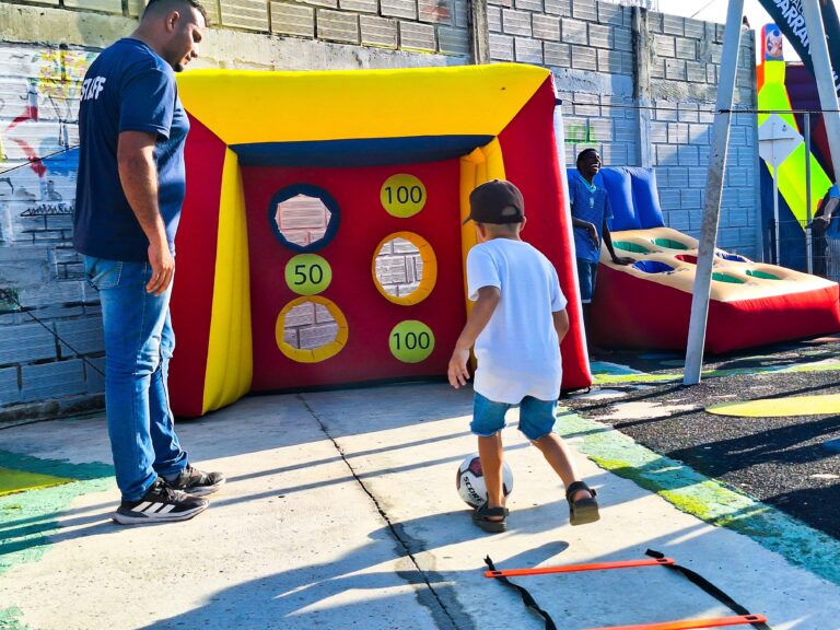 Niño patea un balón hacia un arco inflable junto a un funcionario de Recreación y Deportes durante jornada recreodeportiva en Barranquilla.