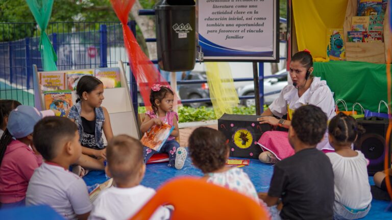 Niños participan en actividad de lectura al aire libre con una cuentacuentos, rodeados de libros y decoraciones coloridas.
