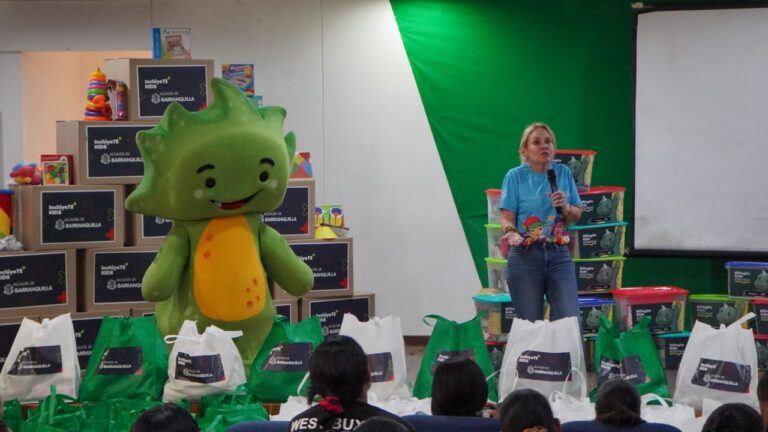 Patricia Vargas habla con micrófono en entrega de kits educativos, acompañada por mascota institucional infantil.