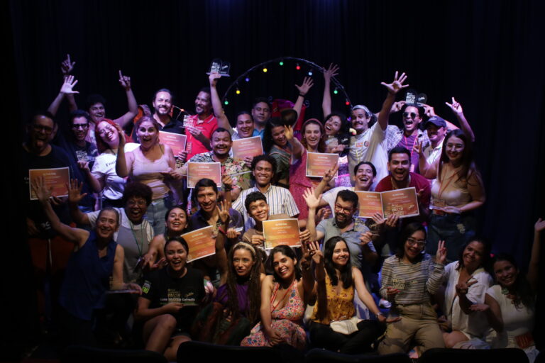 Grupo de artistas celebra con certificados y trofeos sobre escenario tras función.