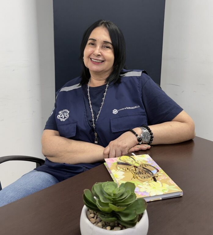 Funcionaria con chaleco institucional, sentada y sonriendo.