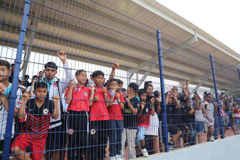 Grupo de niños y jóvenes observan desde detrás de una valla metálica en el polideportivo La Magdalena de Barranquilla.