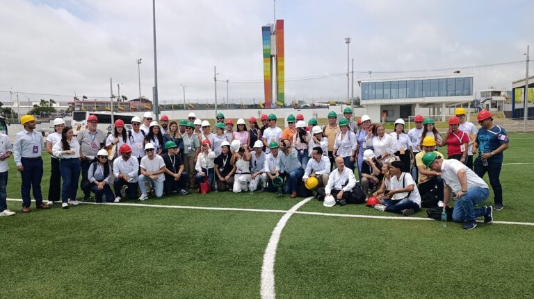 Grupo de asistentes del Foro de Desarrollo Local con cascos de seguridad posa en campo de césped en Tecnoglass, con la Ventana al Mundo de fondo.