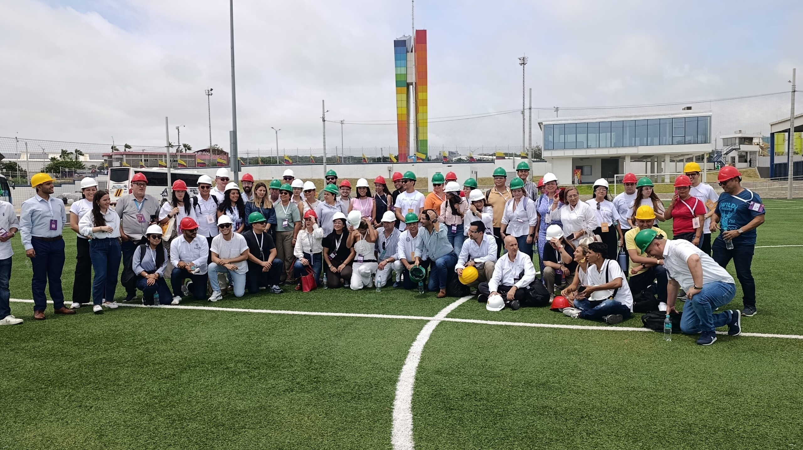 Grupo de asistentes del Foro de Desarrollo Local con cascos de seguridad posa en campo de césped en Tecnoglass, con la Ventana al Mundo de fondo.