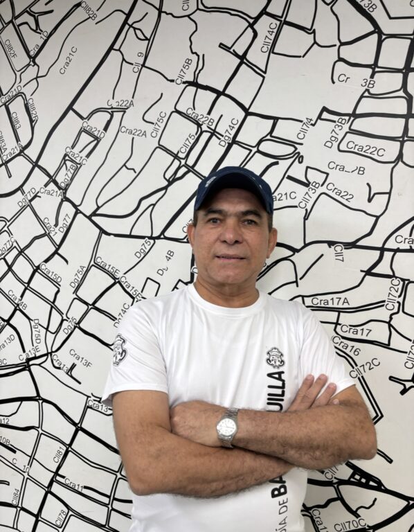 Funcionario posa en plano medio, con camiseta institucional blanca, de fondo a un mapa de barrios.