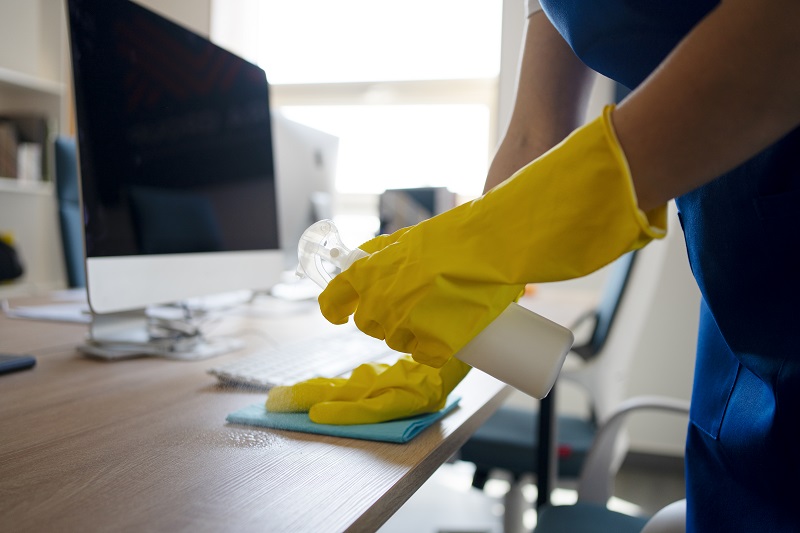 Primer plano de una manos con guantes amarillos, limpieando y desinfectando un escritorio.