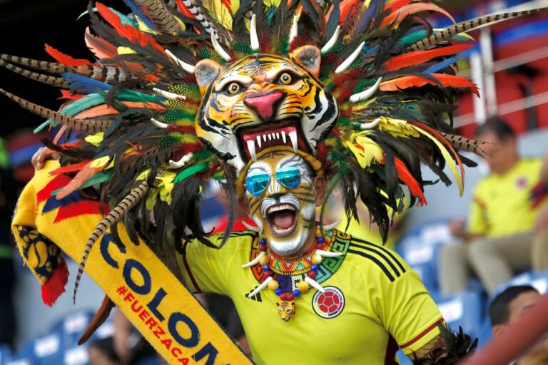 Aficionado colombiano con disfraz de tigre, plumas coloridas y rostro pintado, animando con una bufanda en un estadio.