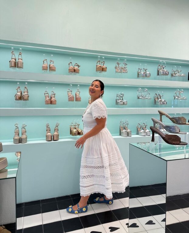 La diseñadora Flor Chaparro posa sonriente en una boutique de calzado, vestida con un vestido blanco.