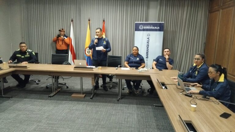 Funcionario de pie con micrófono en mano, da recomendaciones por la temporada de lluvias, junto a otras personas sentadas en mesa de reunión.