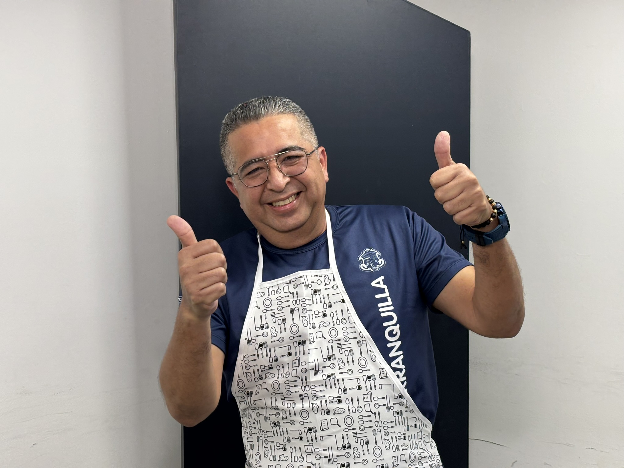 Funcionario posa en plano medio con un sueter institucional y un delantal de cocina, mientras saluda con los dedos pulgares arriba.