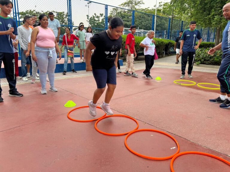 Joven participa en actividad deportiva saltando aros en el suelo, acompañada de un grupo de personas que observan y apoyan.