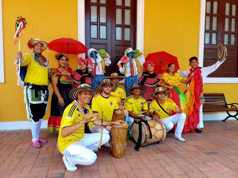 Grupo de bailarines y músicos con trajes típicos del Carnaval de Barranquilla posa frente a una pared amarilla.
