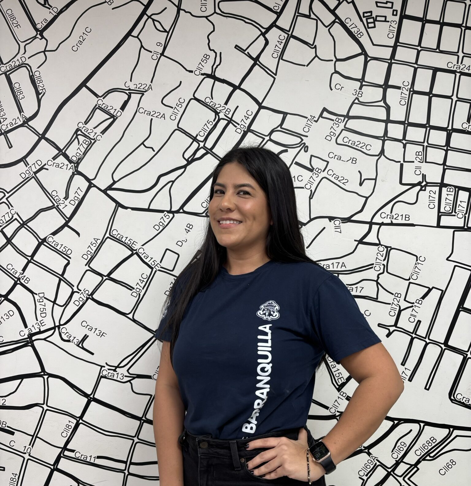 Funcionaria de pie, posando en plano medio con mano en la cintura, vestida de sueter institucional y junto a una pared con mapa de barrial de Barranquilla.