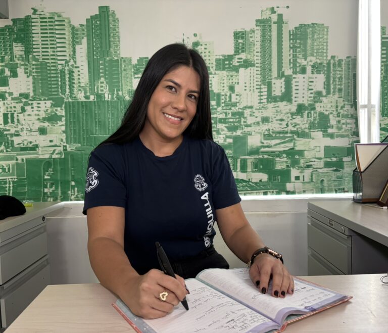 Funcionaria en plano medio horizontal, sentada detras de un escritorio, con un cuaderno abierto y un lapicero en la mano.