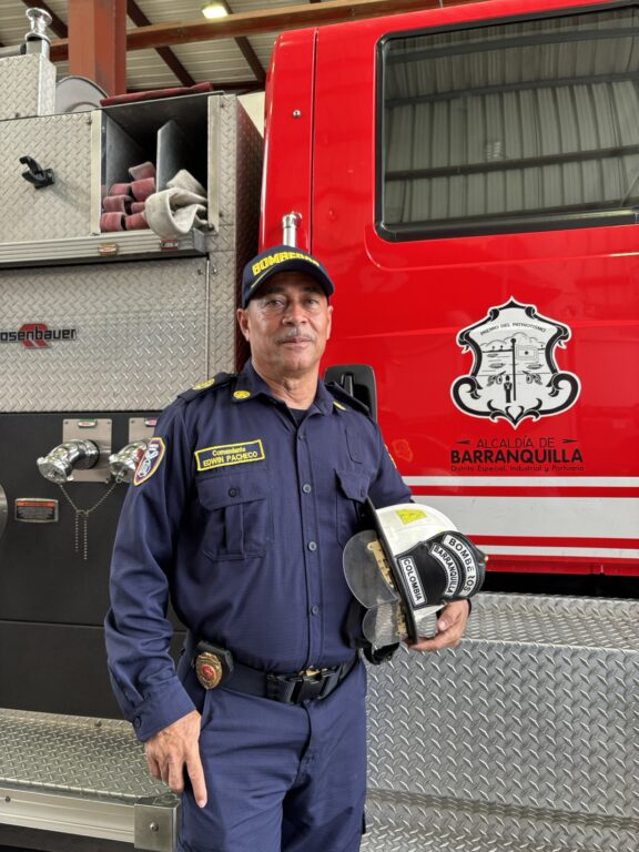 Bombero posa de pie, con el brazo izquierdo sosteniendo su casco y a su espalda el carro de bombero