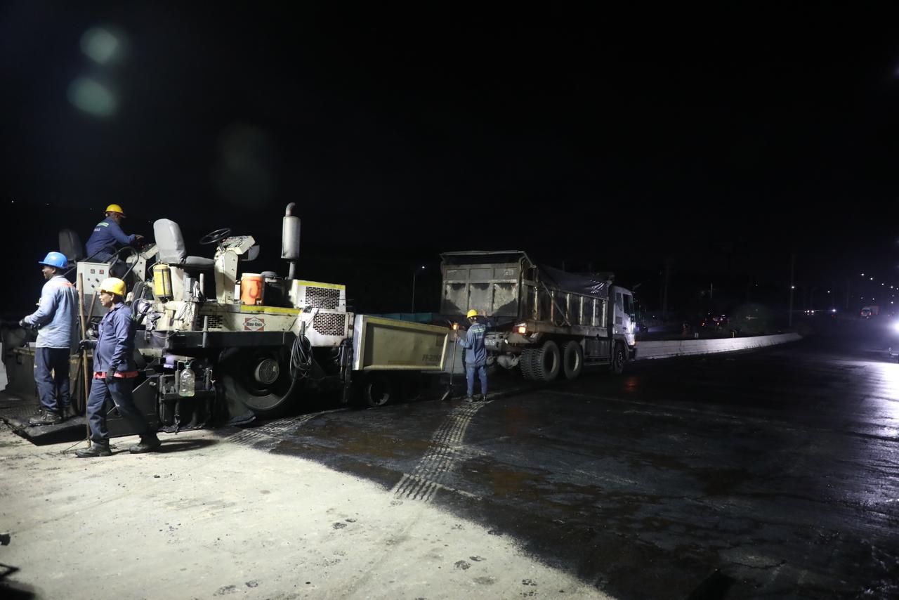 Trabajadores y equipo de volteo en una obra de construcción vial durante la noche