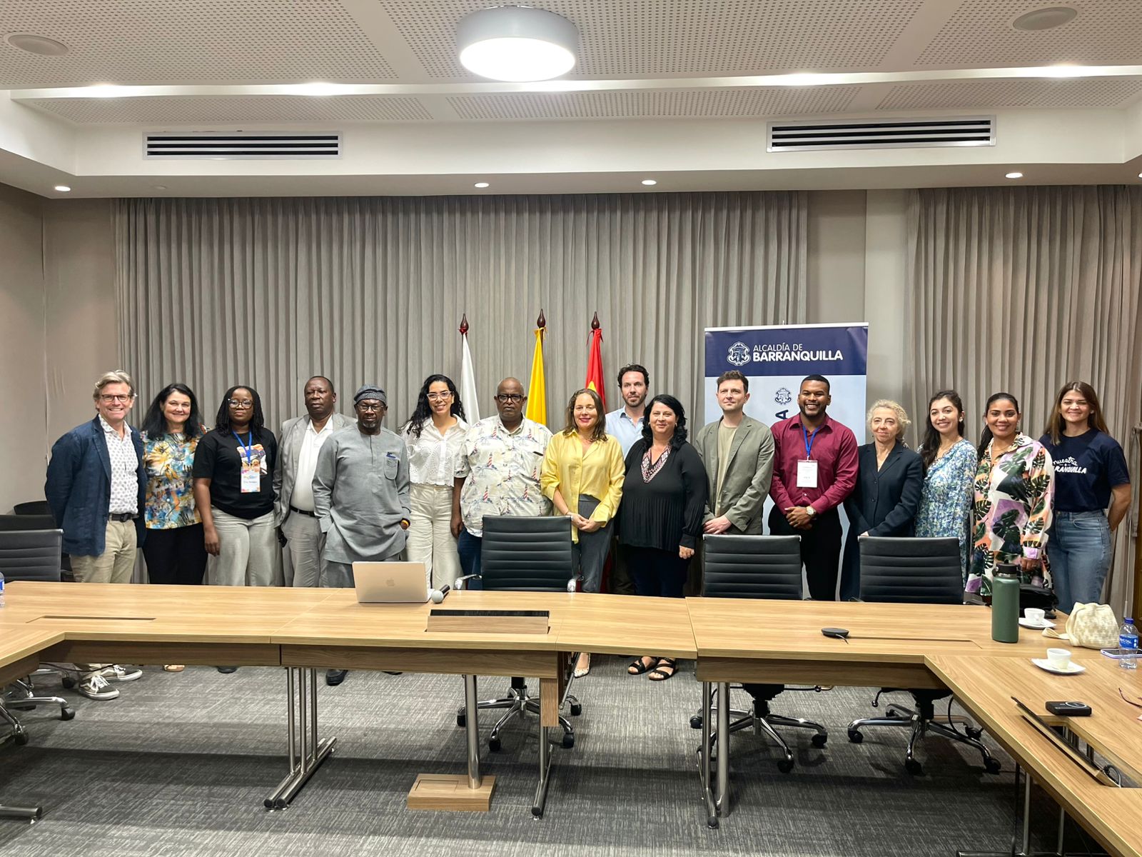 Un grupo de personas de distintas delegaciones internacionales y funcionarios de la Alcaldía de Barranquilla posan de pie en una sala de reuniones. Detrás se observan banderas y un pendón institucional. Todos miran hacia la cámara y sonríen