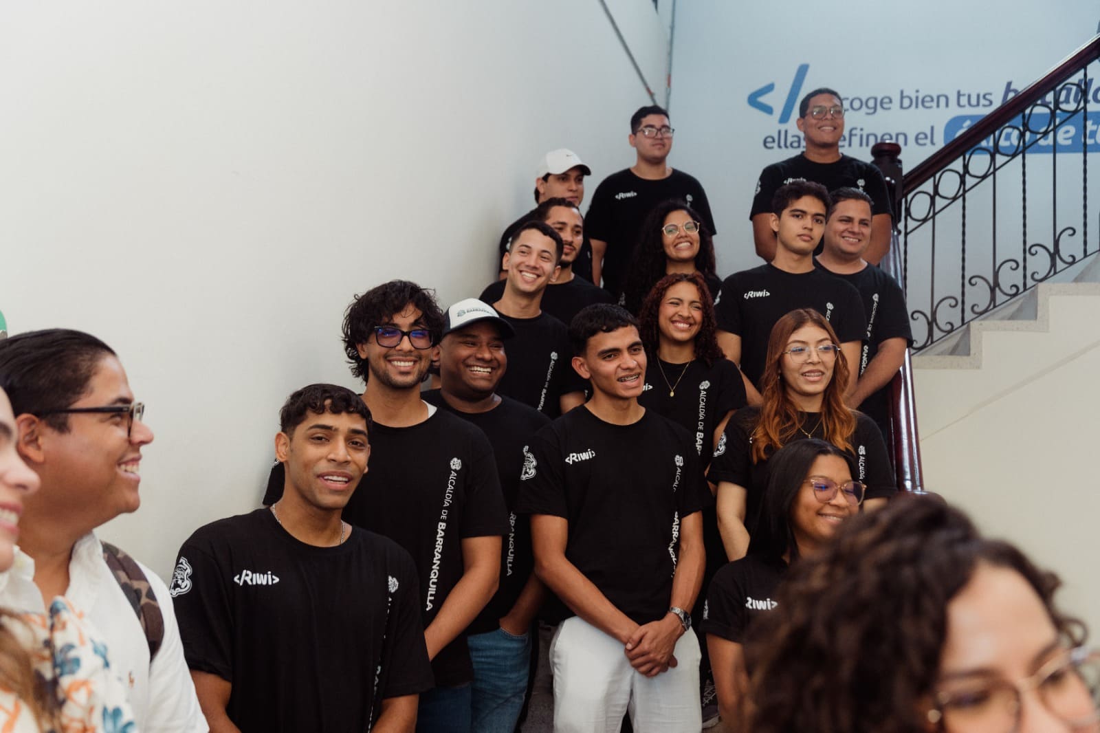 Jóvenes participantes del programa RIWI posan en una escalera con camisetas institucionales de la Alcaldía de Barranquilla, en un entorno educativo que promueve el desarrollo de habilidades tecnológicas.