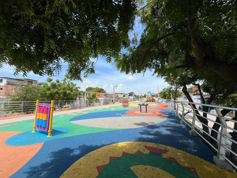 parque infantil al aire libre con un diseño colorido y moderno. El suelo está cubierto con un revestimiento de caucho en tonos vivos como azul, verde, rojo, amarillo y naranja, formando figuras geométricas y decorativas.