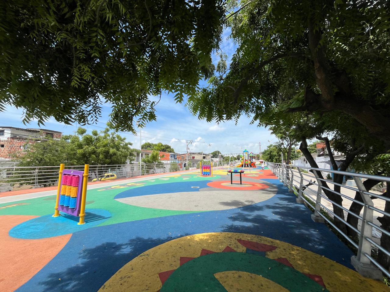 parque infantil al aire libre con un diseño colorido y moderno. El suelo está cubierto con un revestimiento de caucho en tonos vivos como azul, verde, rojo, amarillo y naranja, formando figuras geométricas y decorativas.