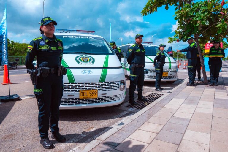 Grupo de agentes de la Policía uniformados, ubicados en formación a un costado de una vía urbana. Detrás de ellos hay vehículos policiales blancos con franjas verdes, estacionados de manera ordenada y con las puertas cerradas.Los policías están de pie, atentos, algunos con las manos al frente o a los costados.