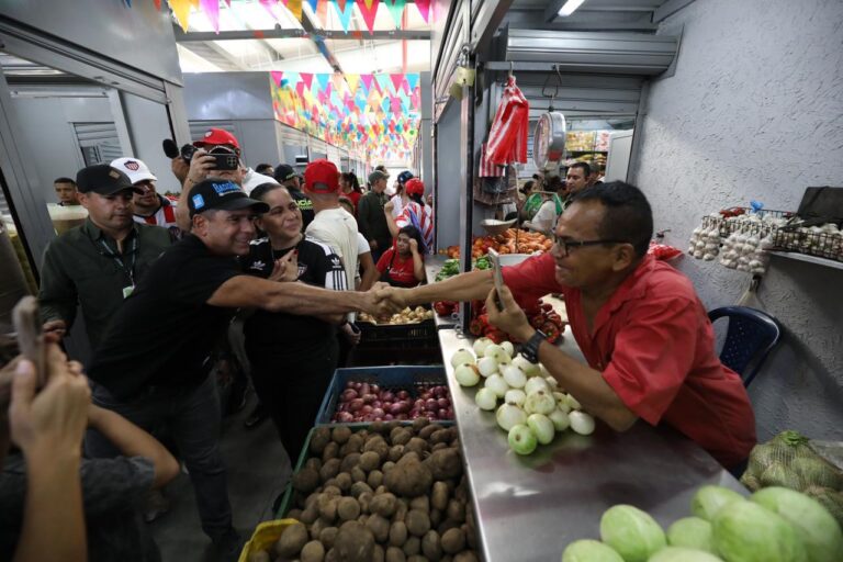 El Acalde Alejandro Char, estrechando la mano a un vendedor. El puesto del vendedor está lleno de productos frescos como cebollas, papas, tomates, ajos y otras verduras.