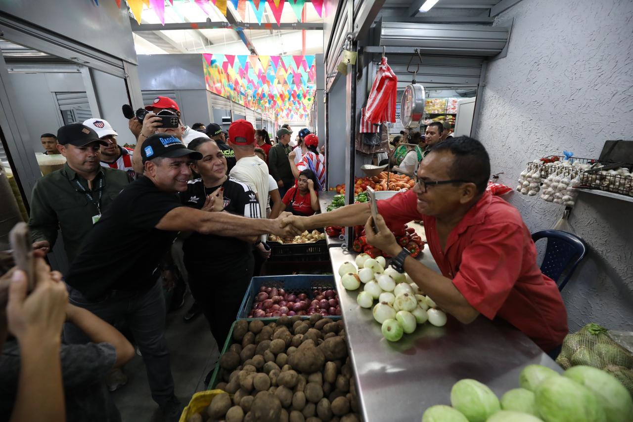 El Acalde Alejandro Char, estrechando la mano a un vendedor. El puesto del vendedor está lleno de productos frescos como cebollas, papas, tomates, ajos y otras verduras.