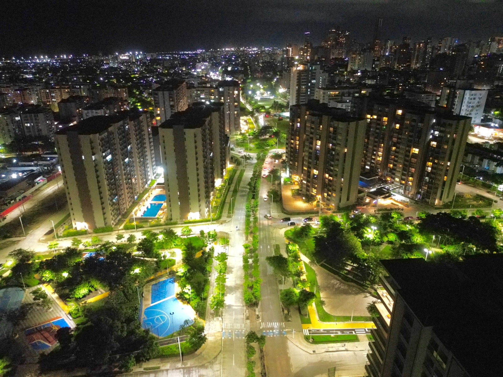 vista aérea nocturna de una zona urbana moderna. Se observan varios edificios altos, probablemente residenciales, iluminados con luces cálidas en sus ventanas. Entre los edificios hay calles amplias y bien organizadas, con árboles y áreas verdes que también están iluminadas por farolas.