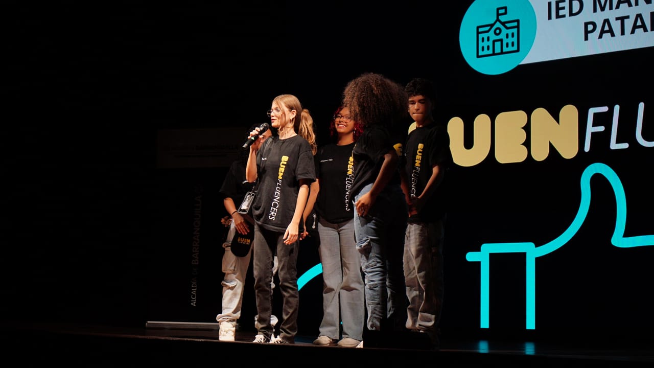 Jovenes durante una presentación del programa BUENFLUENCERS, con una pantalla de fondo que muestra el nombre del proyecto y una referencia a una institución educativa.