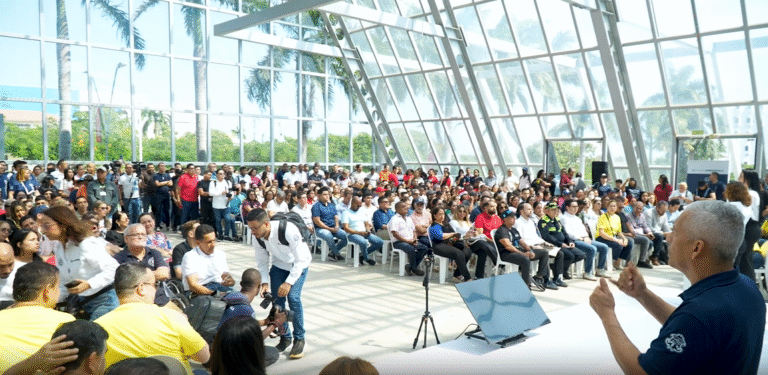 Evento masivo de personas sentadas en filas de sillas plásticas blancas, formando una audiencia numerosa.