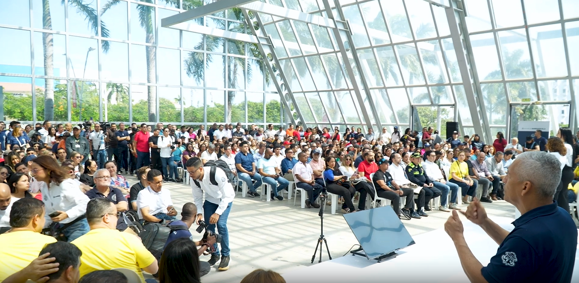 Evento masivo de personas sentadas en filas de sillas plásticas blancas, formando una audiencia numerosa.